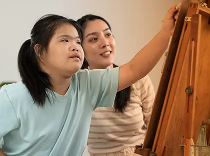 Therapist supports a young girl as she paints on an easel during an ABA therapy session focused on creativity and skill-building