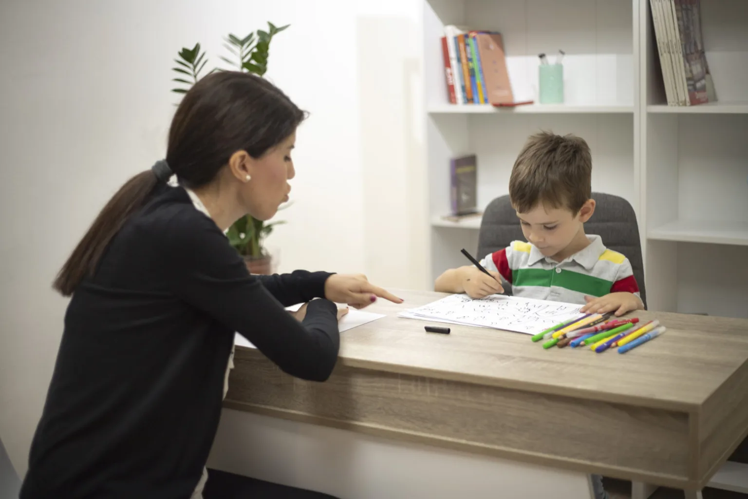 teacher sitting at a desk with a young boy doing writing exercises