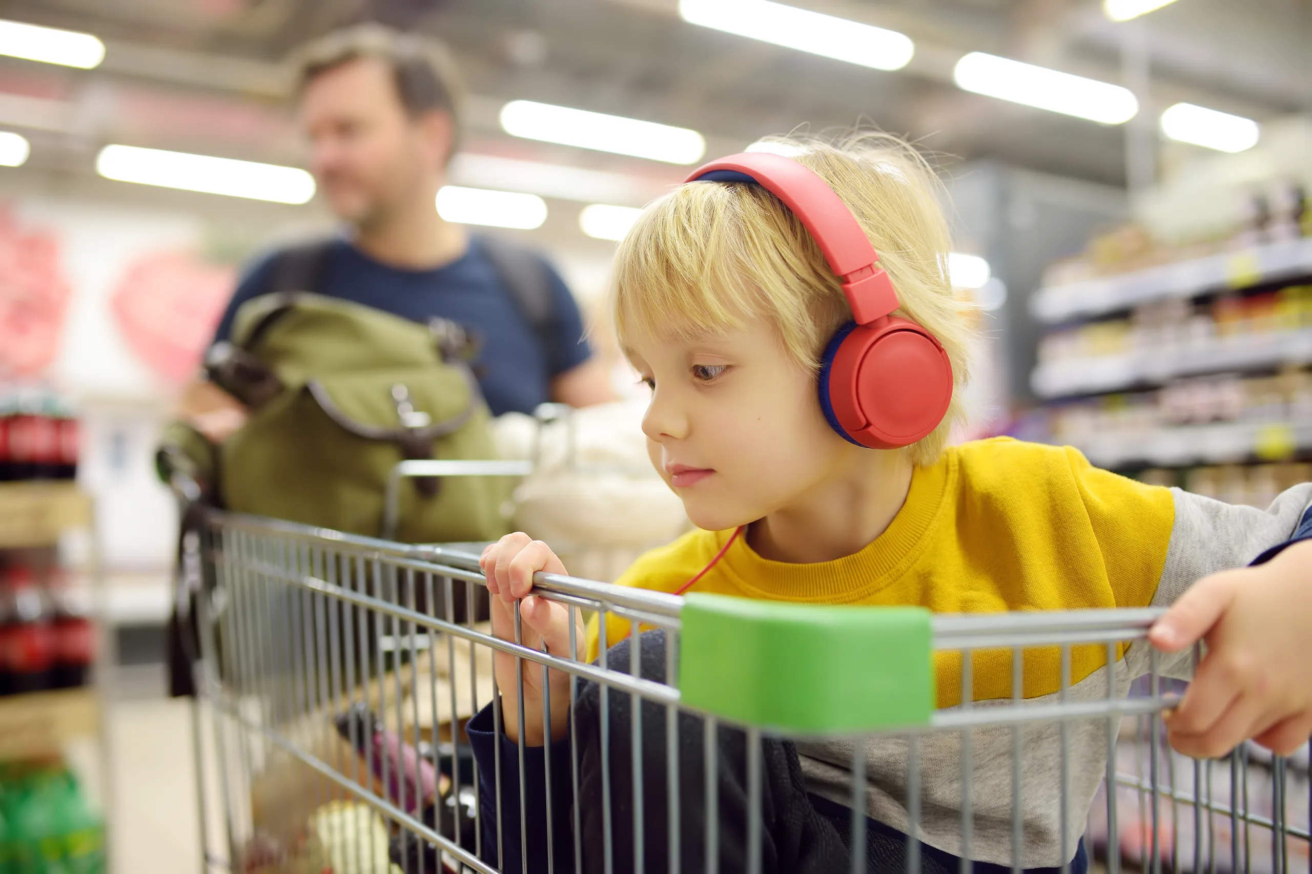 Cute preschooler boy with headphone and player is sitting in a shopping cart at a food store or supermarket. A child is listening to music or an audiobook while his parents buying groceries.