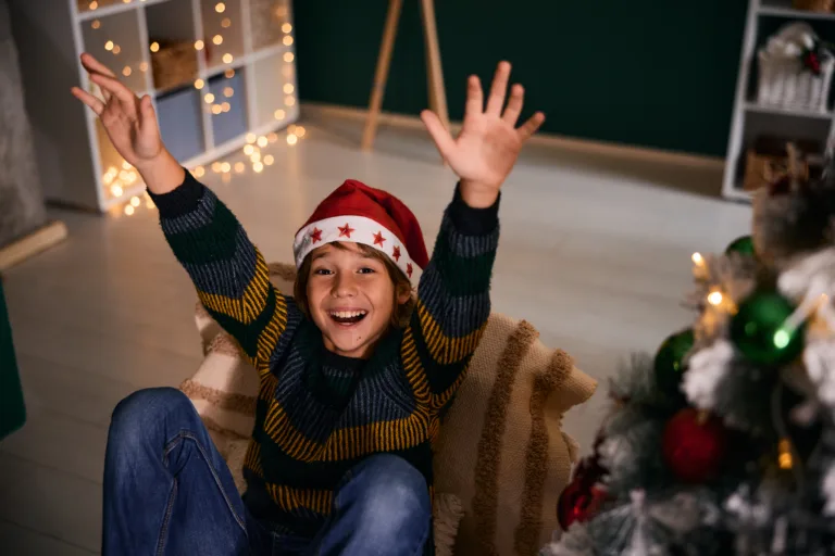 Smiling boy wearing a Santa hat, raising his arms in excitement while sitting near a beautifully decorated Christmas tree at home during the festive holiday season