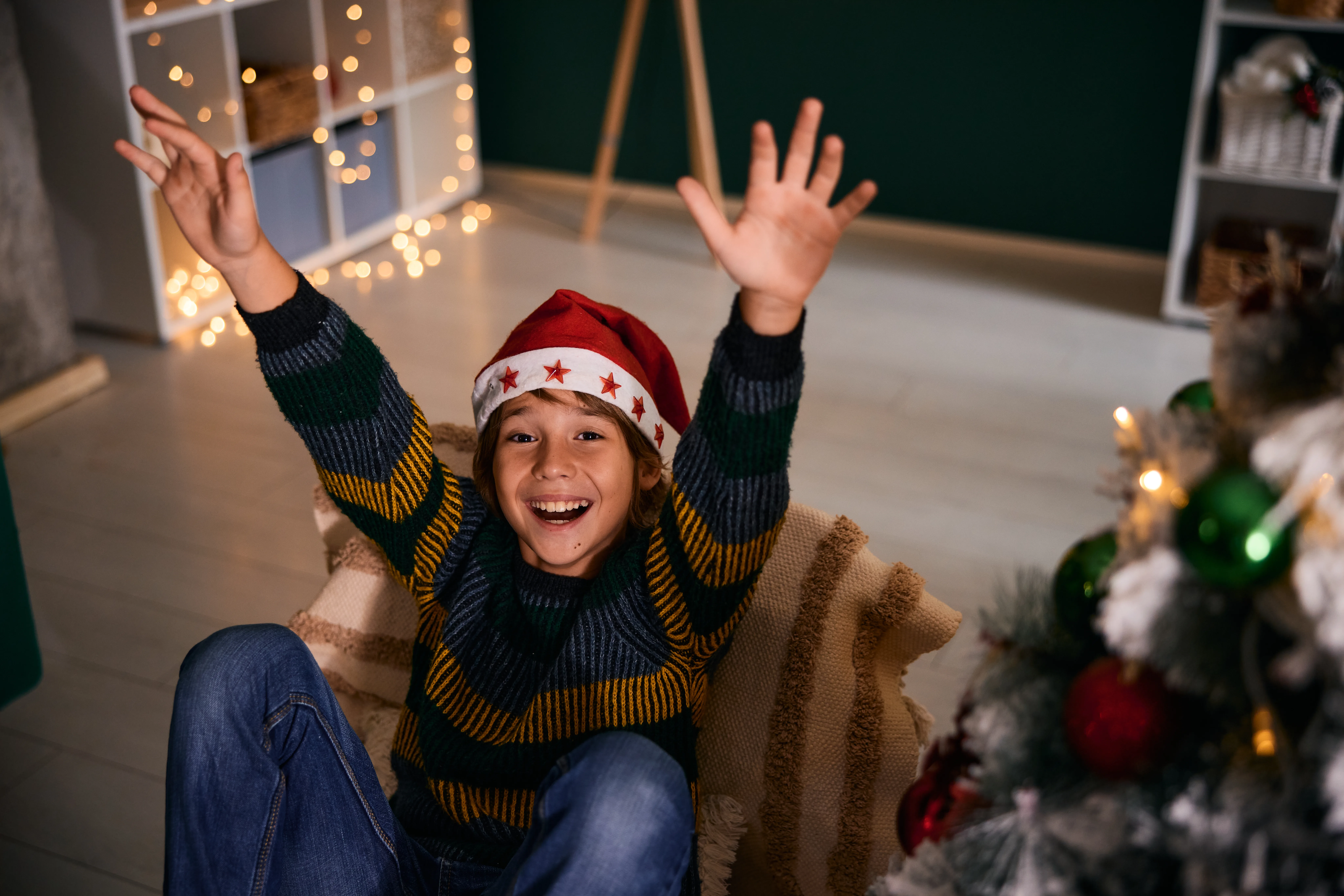 Smiling boy wearing a Santa hat, raising his arms in excitement while sitting near a beautifully decorated Christmas tree at home during the festive holiday season