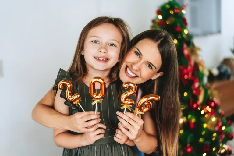 Happy mother and daughter smiling while holding golden balloons forming the number 2026 at home. Decorated Christmas tree in the background adds a festive atmosphere. Concept of family love, new beginnings, and joyful holiday celebration