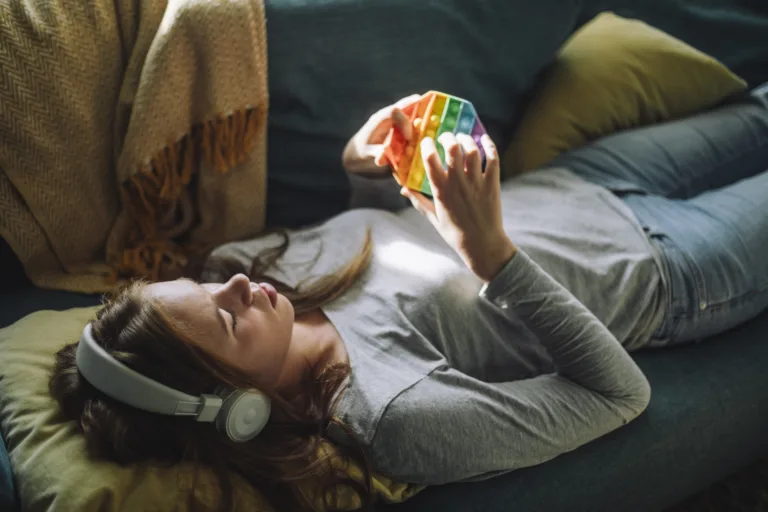 Teenage girl listening to music and playing with push pop toy while lying down on sofa at home