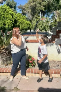 A Board Certified Behavior Analyst (BCBA) and a child stand on a backyard patio, facing each other and raising their hands to complete a high-five.